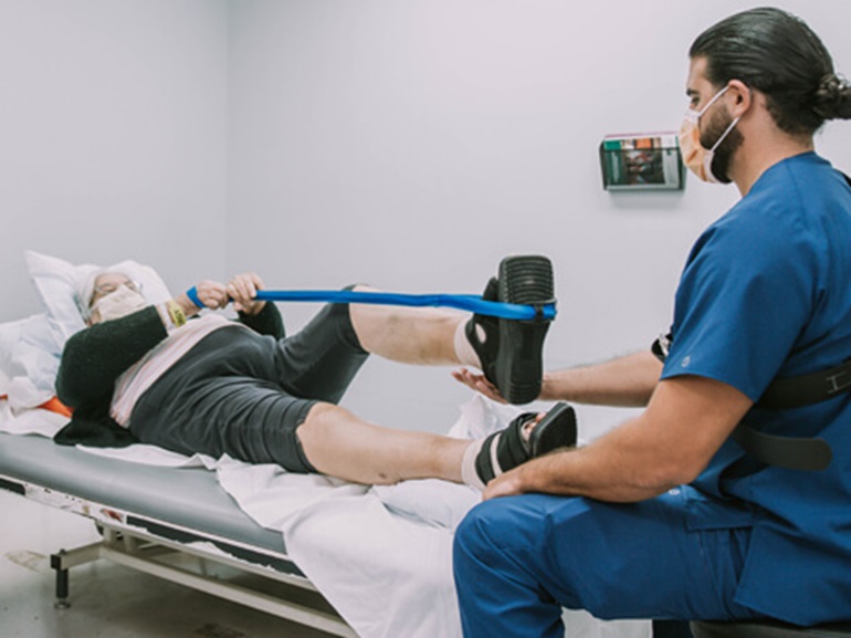 Female patient lying on therapy table holding a resistance band looped around foot to stretch out leg.