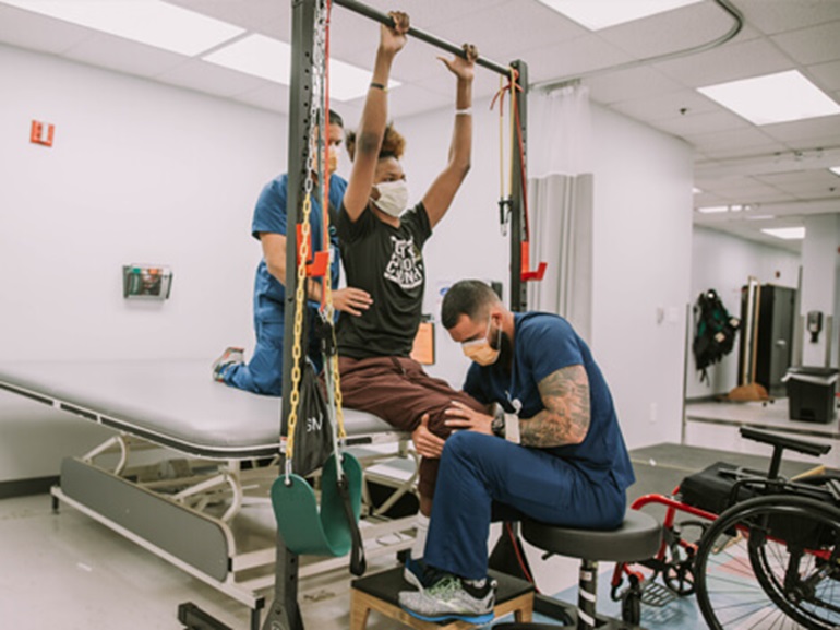 Two therapists working with female patient with raised arms to grab pull-up bar.