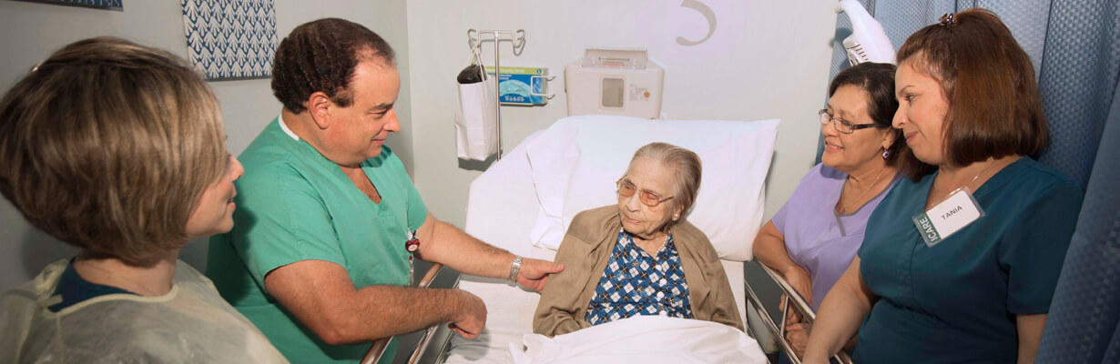 Nurses and male doctor in green scrubs at bedside of older female patient.