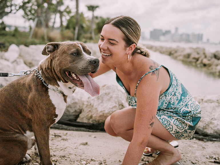 Monica on a beach kneeling down to pet a brown dog.