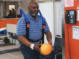 Male patient wearing weight-supported safety harness holding small orange ball.