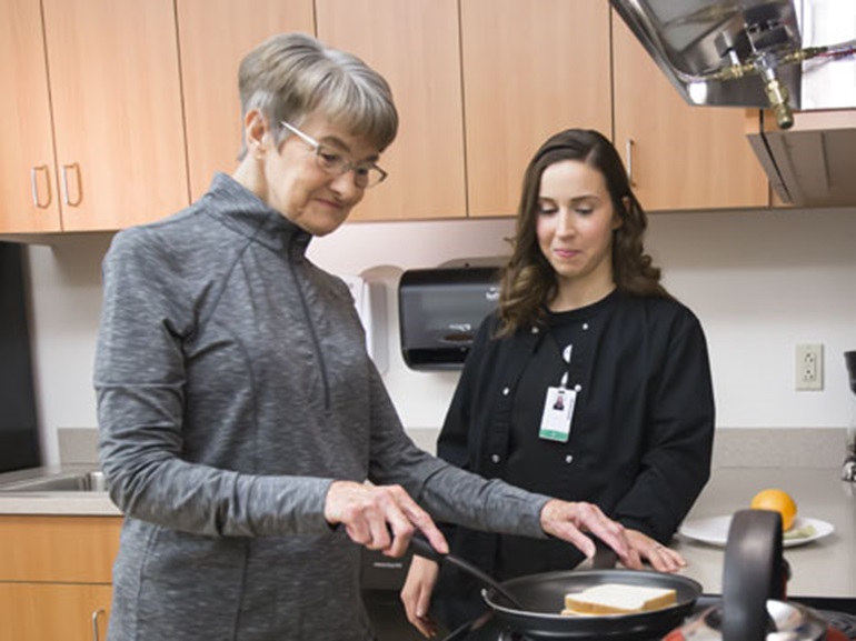 Older female practicing cooking on a stovetop with help of therapist.