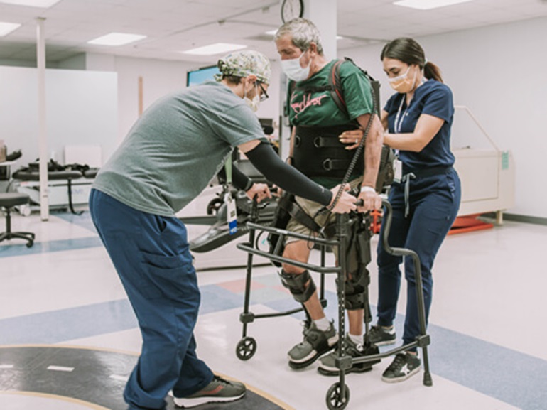 Two therapists helping male patient use a rolling walker in a therapy gym.