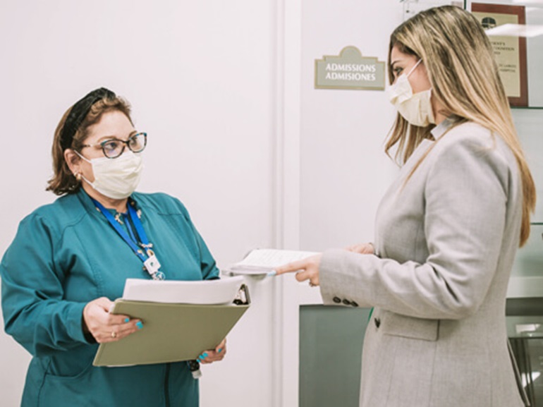 Nurse in green scrubs with mask over nose and mouth talking to a female colleague.