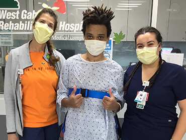 Gusto wearing a hospital gown and yellow mask standing between two thereapists.